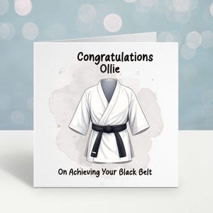 May include: A white karate gi with a black belt is depicted on a greeting card. The card says "Congratulations Ollie On Achieving Your Black Belt" in black text. The background has a watercolour effect.