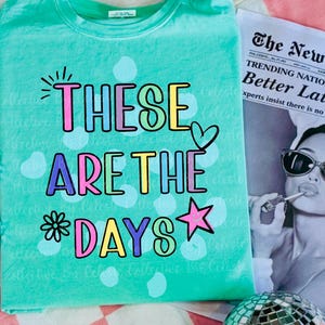 May include: A turquoise t-shirt with the phrase "THESE ARE THE DAYS" in colorful letters and a heart and star graphic. The shirt is next to a newspaper and a disco ball.