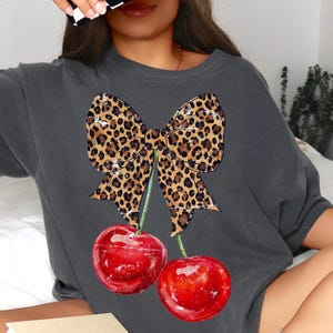 May include: A gray t-shirt with a leopard print bow and two red cherries. The bow is tied with a green stem and the cherries have green stems.