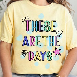 May include: A light yellow t-shirt with the phrase "THESE ARE THE DAYS" in colorful bubble letters. The design includes a heart, star, and flower. The shirt is a casual, comfortable style.