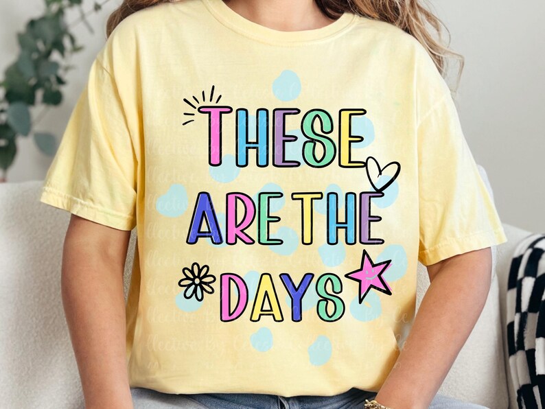 May include: A light yellow t-shirt with the phrase "THESE ARE THE DAYS" in colorful bubble letters. The design includes a heart, star, and flower. The shirt is a casual, comfortable style.