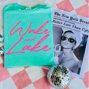 May include: A turquoise t-shirt with the words "Wake and Lake" in a pink and yellow script. A newspaper with the headline "Better Late Than Ugly" and a disco ball are also in the image.