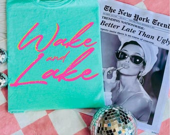 May include: A turquoise t-shirt with the words "Wake and Lake" in a pink and yellow script. A newspaper with the headline "Better Late Than Ugly" and a disco ball are also in the image.