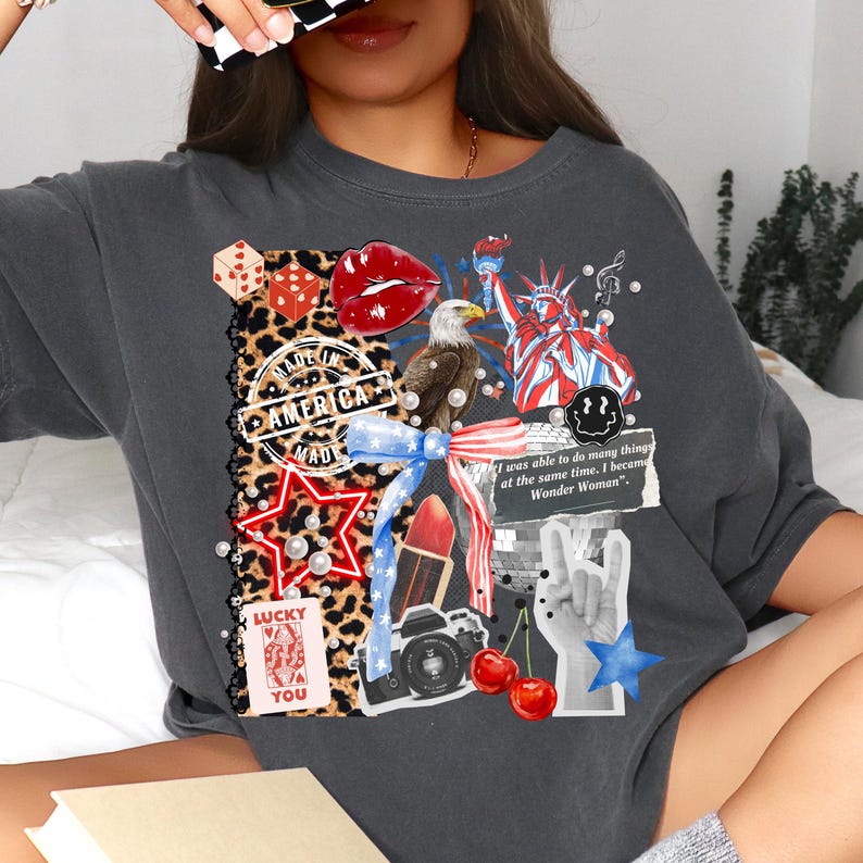 May include: Dark gray t-shirt with a collage design featuring a leopard print, red lips, an eagle, the Statue of Liberty, and the text "MADE IN AMERICA MAD". Other elements include a star, dice, cherries, and the text "LUCKY YOU".