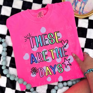 May include: A bright pink sweatshirt with the phrase "THESE ARE THE DAYS" in colorful bubble letters. The design includes a star, heart, and flower. The shirt is folded on a black and white checkered surface.