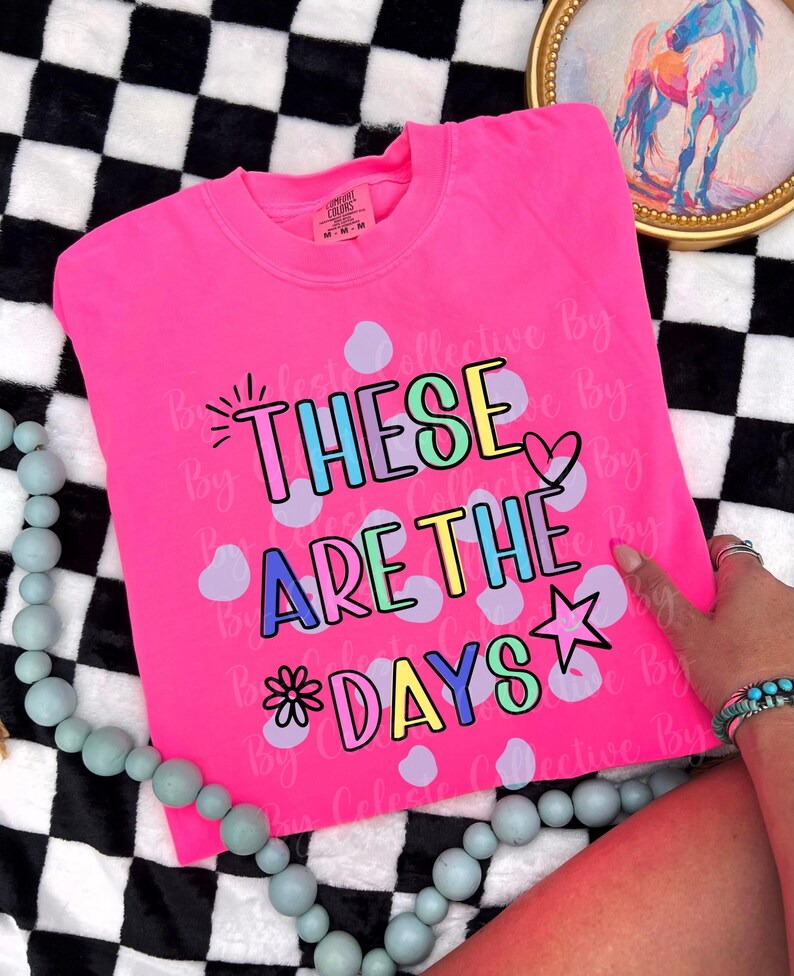 May include: A bright pink sweatshirt with the phrase "THESE ARE THE DAYS" in colorful bubble letters. The design includes a star, heart, and flower. The shirt is folded on a black and white checkered surface.