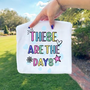 May include: White sweatshirt with the phrase "THESE ARE THE DAYS" in colorful bubble letters. The design includes a heart, star, and floral accents. Light blue polka dots are scattered around the text.
