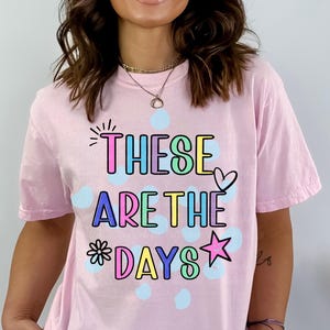May include: A light pink t-shirt with the phrase "THESE ARE THE DAYS" in colorful, outlined letters. The design includes a heart, star, and flower. Light blue polka dots are scattered around the text.
