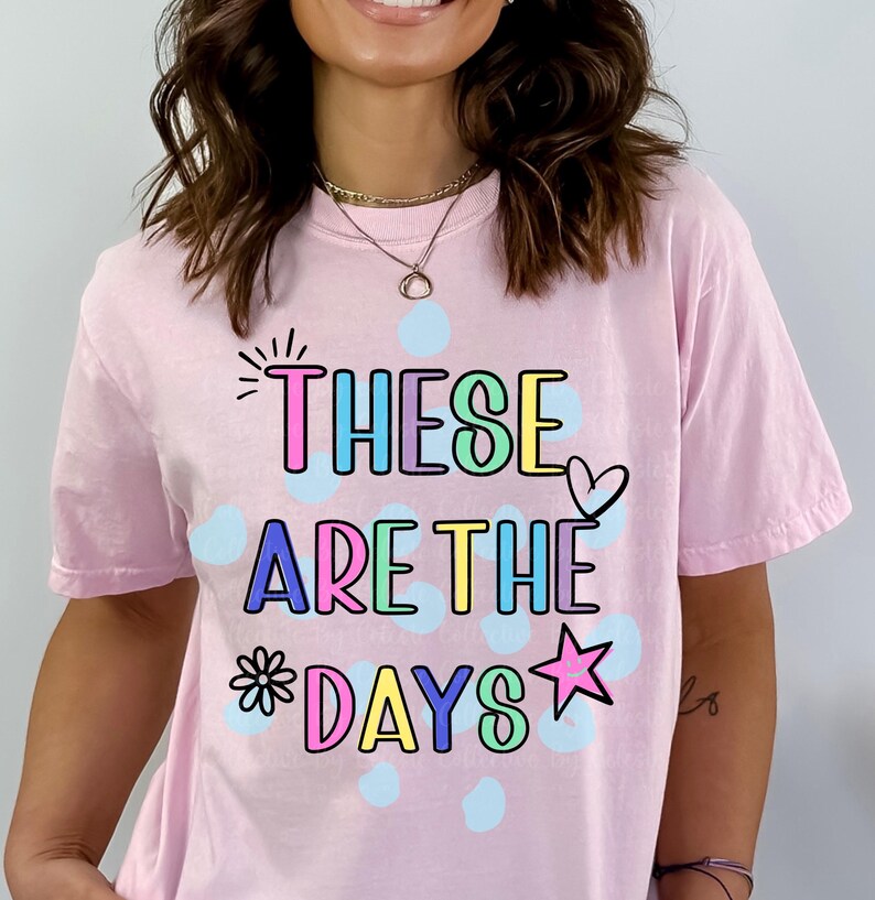 May include: A light pink t-shirt with the phrase "THESE ARE THE DAYS" in colorful, outlined letters. The design includes a heart, star, and flower. Light blue polka dots are scattered around the text.
