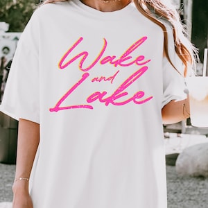 May include: White t-shirt with the text "Wake and Lake" in a vibrant pink and yellow gradient. The text has a slightly distressed, textured appearance, adding a vintage feel to the design. The shirt is a casual, oversized style.