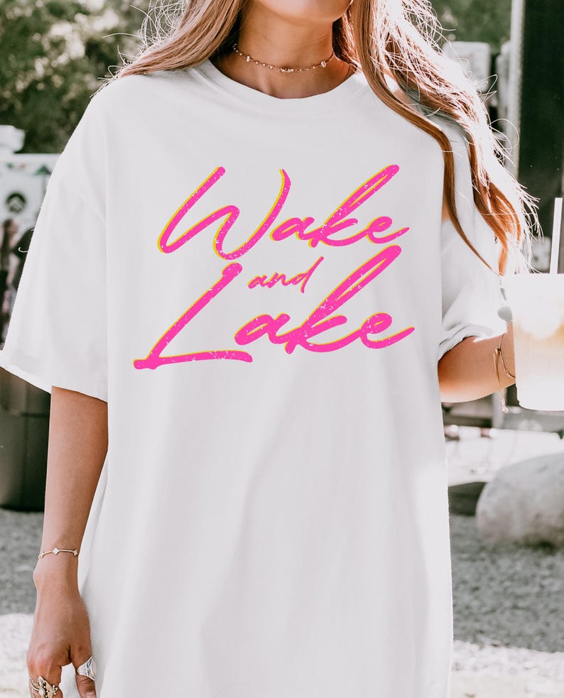 May include: White t-shirt with the text "Wake and Lake" in a vibrant pink and yellow gradient. The text has a slightly distressed, textured appearance, adding a vintage feel to the design. The shirt is a casual, oversized style.
