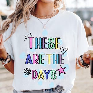 May include: White t-shirt with the phrase "THESE ARE THE DAYS" in colorful bubble letters. The design includes a heart, star, and flower. The shirt is worn by a person wearing sunglasses and jewelry.