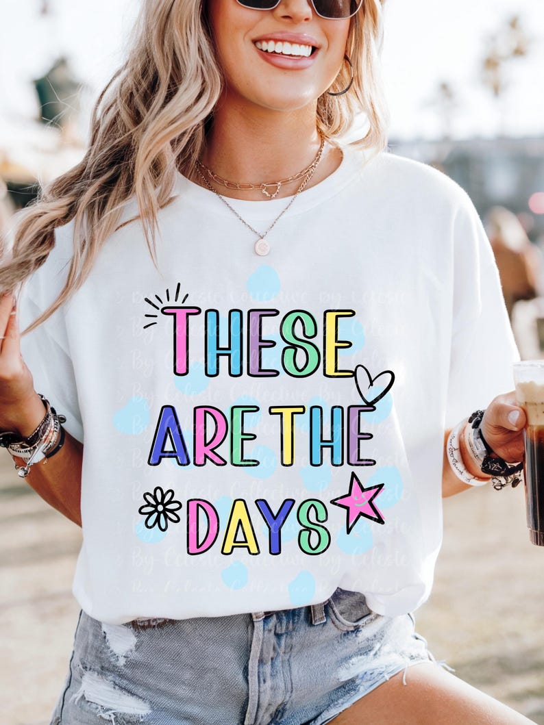 May include: White t-shirt with the phrase "THESE ARE THE DAYS" in colorful bubble letters. The design includes a heart, star, and flower. The shirt is worn by a person wearing sunglasses and jewelry.