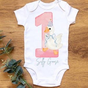 May include: A white onesie with a pink number one and a cartoon goose wearing a party hat. The text "Silly Goose" is printed below the number.