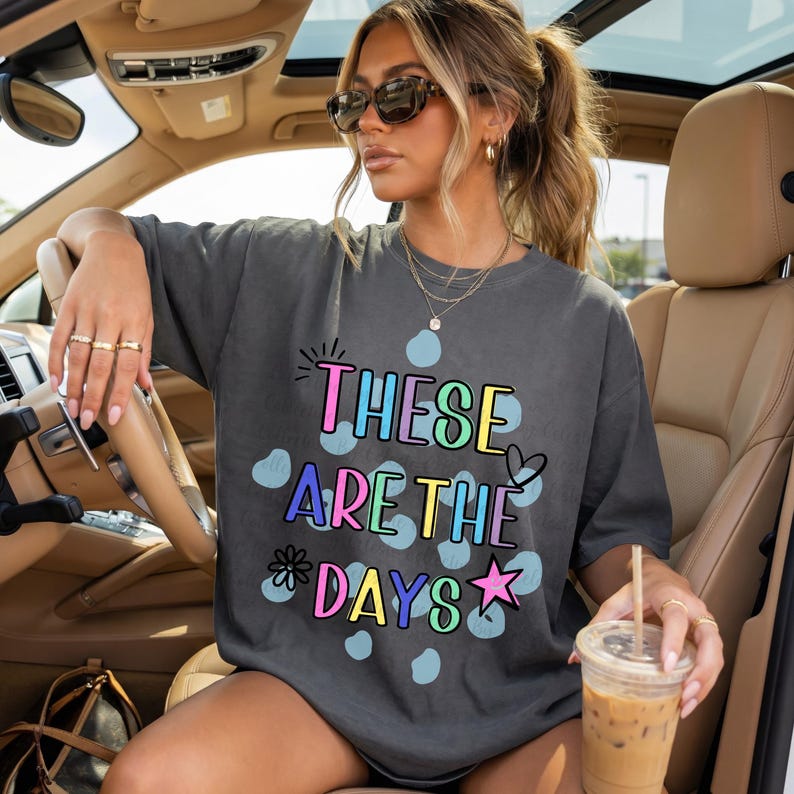 May include: Dark gray t-shirt with the text "THESE ARE THE DAYS" in colorful bubble letters. The shirt has a relaxed fit and is worn by a person in a car. The design includes blue polka dots, a heart, a star, and a flower.