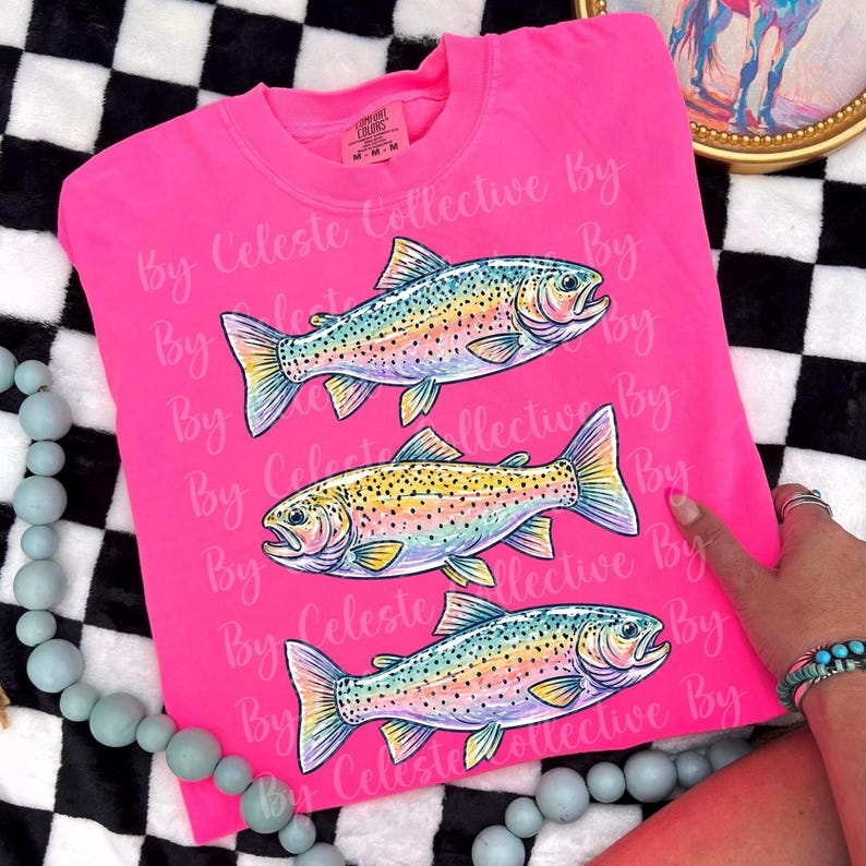 May include: A hot pink t-shirt featuring three colorful fish illustrations. The fish have rainbow-colored bodies with yellow, blue, and green accents. The text "By Celeste Collective By" is printed repeatedly on the shirt.