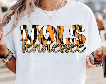 May include: White t-shirt with the text "VOLS Tennessee" in orange, black, and gold glitter lettering. The shirt has a crew neck and short sleeves. The design features a tiger stripe pattern.