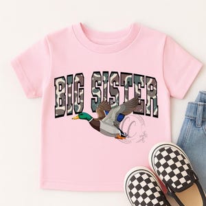May include: A light pink t-shirt featuring the text "BIG SISTER" in a camouflage design. A cartoon duck in flight is below the text. Checkered black and white slip-on shoes and denim jeans are visible.