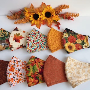May include: Ten different fabric face masks with various autumn-themed patterns, including sunflowers, leaves, and floral prints. The masks are all in a variety of colours, including brown, orange, yellow, and white.