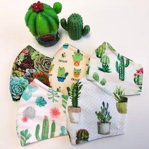 Puede incluir: Cinco mascarillas de tela diferentes con diseños de cactus y suculentas. Las mascarillas son todas blancas con detalles en verde, rosa y marrón. Las mascarillas están dispuestas en círculo sobre una superficie blanca.