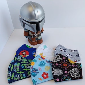 May include: Five different fabric face masks with various Disney and Star Wars characters. The masks are blue, gray, black, white, and light blue. The masks are arranged in a semi-circle on a white surface. A silver and brown Mandalorian figurine is in the background.