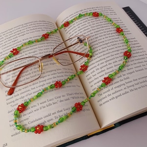 Puede incluir: Primer plano de un libro con un par de gafas y un collar de cuentas rojas y verdes. El collar está hecho de pequeñas cuentas redondas y tiene un patrón floral. El libro está abierto en una página con texto sobre una historia.