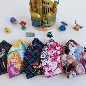 Pode incluir: Cinco máscaras de tecido com diferentes desenhos de personagens da Disney. As máscaras são todas de forma triangular e têm uma variedade de cores e padrões. Os desenhos incluem Úrsula de A Pequena Sereia, Rapunzel de Enrolados, vitrais, Ariel de A Pequena Sereia e personagens de Frozen.