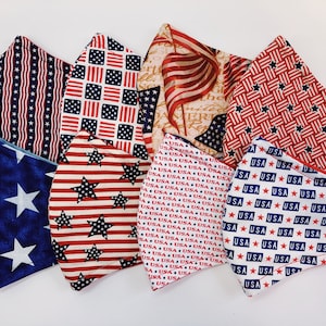 Puede incluir: Una colección de seis mascarillas faciales con varios diseños patrióticos, incluyendo estrellas, rayas y el texto "USA" en rojo, blanco y azul.