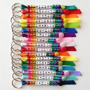 May include: A set of 18 colourful beaded keychains with names spelled out on each one. The beads are rainbow coloured and have glitter embedded in them. Each keychain has a silver key ring and a ribbon attached to the bottom.