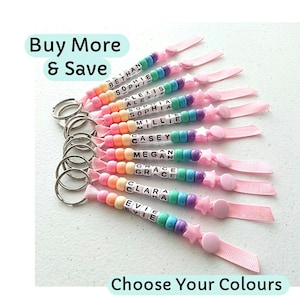 May include: A collection of personalized keychains with rainbow-colored beads spelling out names. Each keychain has a silver ring, a pink ribbon, and a star-shaped charm. The text "Buy More & Save" and "Choose Your Colours" are also visible.