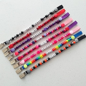 May include: A selection of beaded dummy clips in assorted colours. Each clip has a name spelled out with white letter beads. The clips have a metal clasp and a ribbon loop with a button. The beads are in shades of red, purple, pink, and rainbow.