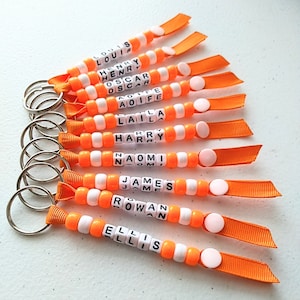 May include: A collection of personalized keychains with names spelled out in white letter beads. The keychains feature orange and white beads, orange ribbon, and silver key rings. Each keychain has a different name, such as "LOUIS" and "ELLIS".