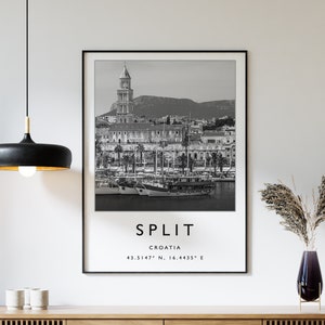 May include: Black and white photograph of the city of Split, Croatia, with the text "SPLIT, CROATIA, 43.5147° N, 16.4435° E" printed below the image. The photograph is framed and hanging on a white wall above a wooden cabinet with slatted doors.