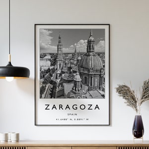 May include: Black and white photograph of the city of Zaragoza, Spain. The photo shows a cityscape with several buildings, including a large church with a dome. The text "ZARAGOZA" is printed in bold, black letters at the bottom of the photo, with the words "SPAIN" and "41.6489° N, 0.8891° W" printed below.