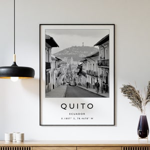 May include: Black and white photograph of a street scene in Quito, Ecuador. The photo shows a street lined with buildings, with a hill in the background. The text "QUITO" and "ECUADOR" are printed below the image, along with the coordinates "0.1807&deg; S, 78.4678&deg; W".