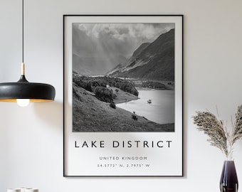 May include: Black and white photograph of a lake and mountains in the Lake District, United Kingdom. The text "LAKE DISTRICT", "UNITED KINGDOM", and "54.5772° N, 2.7975° W" is printed below the image.