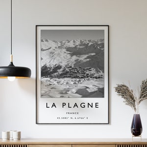 Könnte beinhalten: Schwarzweißfoto einer schneebedeckten Bergkette mit einem kleinen Dorf im Tal. Der Druck ist schwarz gerahmt und trägt den Text "LA PLAGNE, FRANKREICH, 45.5082° N, 6.6766° E" unter dem Bild.