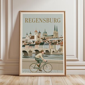 以下が含まれることがあります： 金色の文字でREGENSBURGと書かれた都市のイラストが描かれた額装アートプリント。石造りの橋と歴史的な建物が並ぶ川沿いを自転車で走る人物が描かれています。