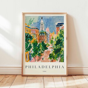 May include: A framed art print of Philadelphia, USA, with a vibrant watercolor illustration of a city street. The artwork depicts buildings in orange, yellow, and blue, with green trees along the street. The word "PHILADELPHIA" is printed at the bottom of the print.