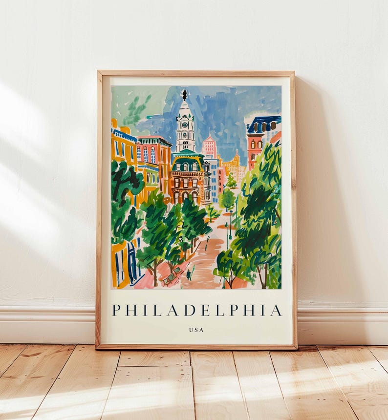May include: A framed art print of Philadelphia, USA, with a vibrant watercolor illustration of a city street. The artwork depicts buildings in orange, yellow, and blue, with green trees along the street. The word "PHILADELPHIA" is printed at the bottom of the print.