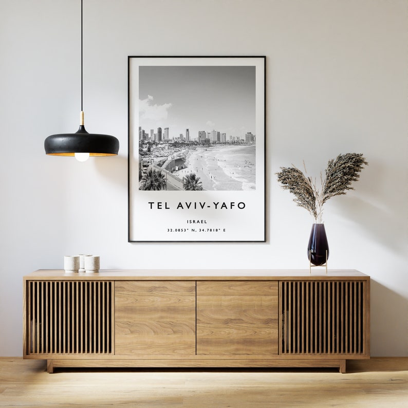 May include: Black and white photograph of the Tel Aviv-Yafo skyline in Israel. The photo is framed in a black frame and hangs above a wooden cabinet with a vase of dried flowers on top.