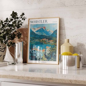 May include: A framed vintage travel poster with the text "WHISTLER CANADA" and a depiction of a mountain range and lake. The poster is on a countertop with a silver pitcher, a wooden cutting board, a ceramic vase, and a silver bowl with lemons.