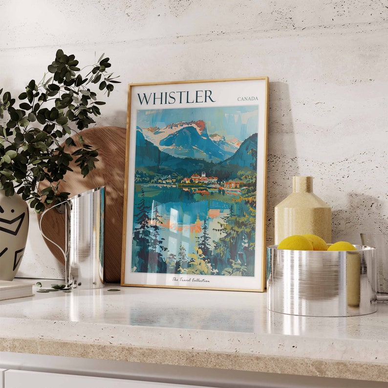 May include: A framed vintage travel poster with the text "WHISTLER CANADA" and a depiction of a mountain range and lake. The poster is on a countertop with a silver pitcher, a wooden cutting board, a ceramic vase, and a silver bowl with lemons.