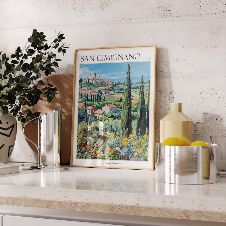 May include: A framed vintage travel poster with a gold frame. The poster features a colorful illustration of the Italian town of San Gimignano, with rolling hills and cypress trees. The text on the poster reads "San Gimignano, Italy, The Travel Collection."