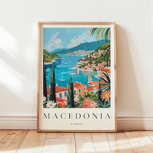 Könnte beinhalten: Ein gerahmtes Reiseplakat für Mazedonien, Europa. Das Kunstwerk zeigt eine Küstenstadt mit rotbedachten Gebäuden, Segelbooten auf dem türkisfarbenen Wasser und grünen Hügeln. Das Plakat trägt das Wort "MACEDONIA" in großen Buchstaben.