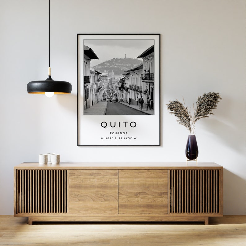 May include: Black and white photograph of a street scene in Quito, Ecuador. The photo shows a street lined with buildings, with a hill in the background. The text "QUITO" and "ECUADOR" are printed below the image, along with the coordinates "0.1807&deg; S, 78.4678&deg; W".