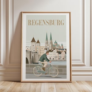 以下が含まれることがあります： 金色の文字でREGENSBURGと書かれた額装された旅行ポスター。イラストには、石橋の上で水色の自転車に乗る人物と、ドイツのレーゲンスブルクの街並み、大聖堂、川が描かれています。