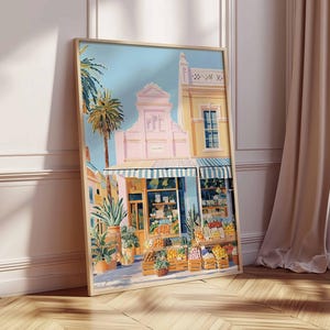 May include: A framed art print showing a sunny street scene with a fruit stand. The artwork features a building with a pink facade, a blue awning, and palm trees. Crates of produce are displayed in front of the shop, with a light blue sky in the background.