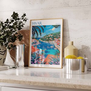 Puede incluir: Un p&oacute;ster enmarcado con una ilustraci&oacute;n colorida de un pueblo portuario con el texto "HVAR CROATIA The Travel Collection". El p&oacute;ster est&aacute; en una encimera con una jarra plateada, un jarr&oacute;n y un cuenco de limones.