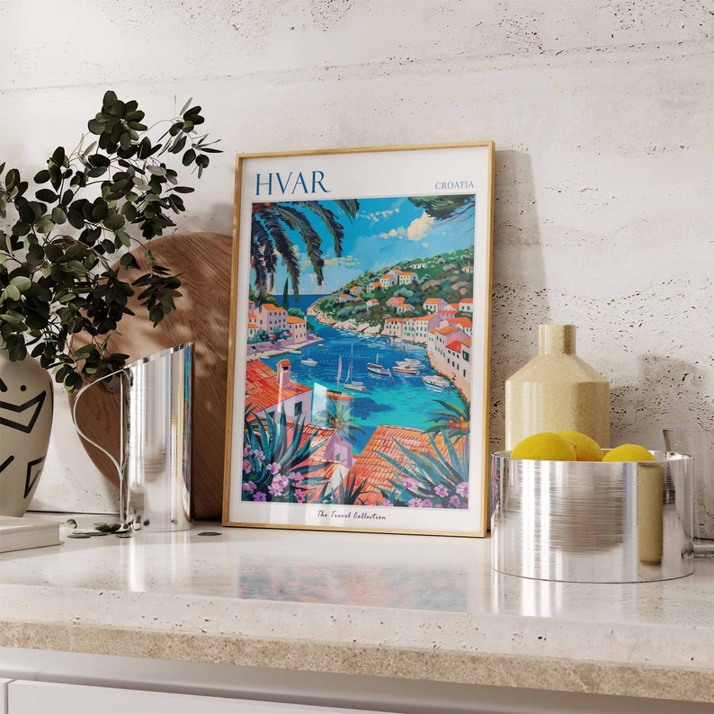 Puede incluir: Un p&oacute;ster enmarcado con una ilustraci&oacute;n colorida de un pueblo portuario con el texto "HVAR CROATIA The Travel Collection". El p&oacute;ster est&aacute; en una encimera con una jarra plateada, un jarr&oacute;n y un cuenco de limones.